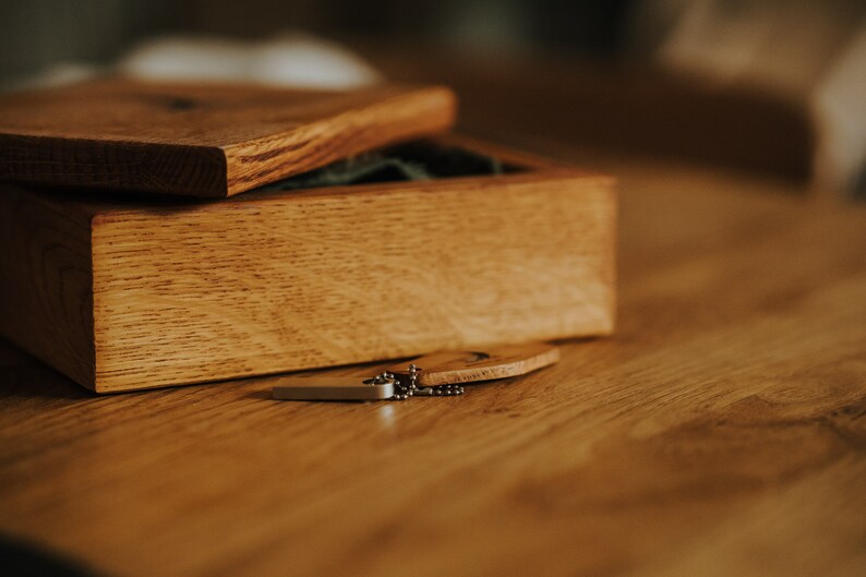 May include: A wooden box with a hinged lid, open to reveal a green interior. A small wooden tag and a silver-colored chain are next to the box. The box and tag are on a wooden surface.