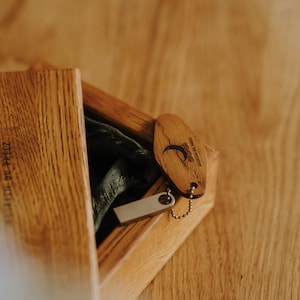 May include: Wooden box with a hinged lid, containing a USB drive and a wooden keychain. The keychain is oval-shaped with a small metal chain and a design. The box and keychain are made of light-colored wood.