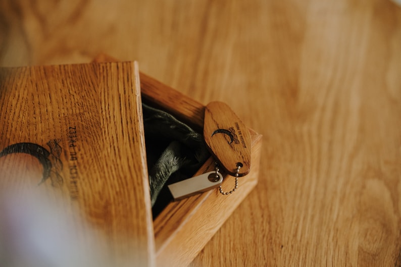 May include: Wooden box with a hinged lid, containing a USB drive and a wooden keychain. The keychain is oval-shaped with a small metal chain and a design. The box and keychain are made of light-colored wood.