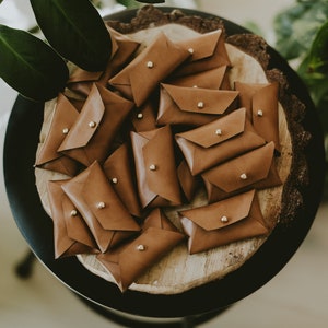 May include: A collection of small, brown leather pouches with a flap closure and a gold-colored stud. The pouches are arranged on a wooden surface, with green foliage in the background. The pouches are likely for small items.