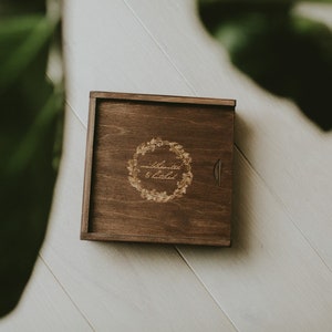 May include: A wooden box with a hinged lid, featuring a laser-engraved wreath design and the text "celebrated & loved".