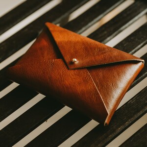 Könnte beinhalten: Eine braune Leder-Kupplung in Briefform mit einem goldenen Knopfverschluss. Die Kupplung liegt auf einer schwarz-weiß gestreiften Oberfläche.