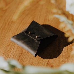 May include: A black leather envelope-style pouch with two brass rivets. The pouch is sitting on a wooden surface.