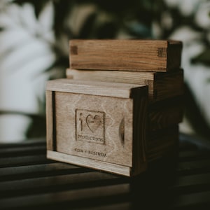 May include: A wooden box with the text "i love productions com + belinda" engraved on the front. The box is stacked on top of other wooden boxes.