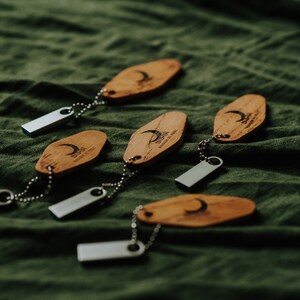 May include: Wooden keychains with a crescent moon design and the text "DUSK" are attached to silver USB drives. The keychains are on a green fabric surface. The keychains are made of wood.
