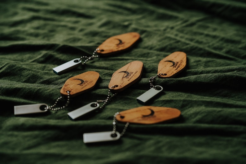May include: Wooden keychains with a crescent moon design and the text "DUSK" are attached to silver USB drives. The keychains are on a green fabric surface. The keychains are made of wood.