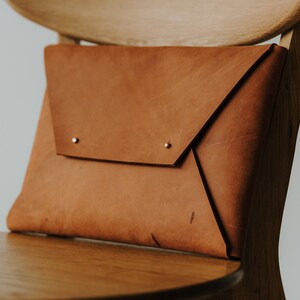 May include: A brown leather envelope-style clutch with a flap closure and two brass rivets. The clutch is sitting on a wooden chair.