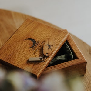 May include: Wooden box with a hinged lid, containing a small wooden tag and a USB drive. The box and tag are engraved with a crescent moon and text. The box is open, revealing a green fabric lining.