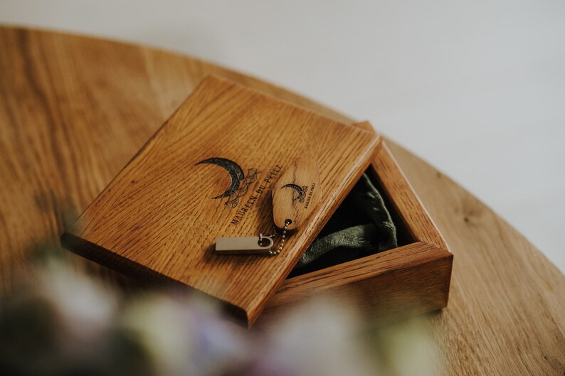 May include: Wooden box with a hinged lid, containing a small wooden tag and a USB drive. The box and tag are engraved with a crescent moon and text. The box is open, revealing a green fabric lining.