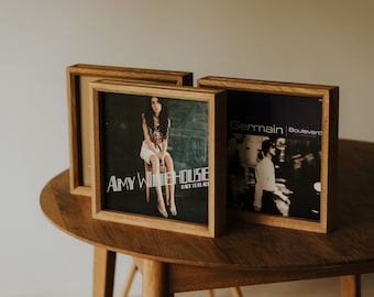 May include: Three wooden frames with black and white album covers. The frames are on a wooden table. The album covers feature Amy Winehouse, Back to Black, and Germain Boulevard.