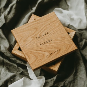 Peut inclure: Boîte en bois clair, gravée des noms "Chiara + Pierre" et de la date "11.09.2021". Posée sur un tissu gris avec un ruban blanc.