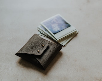 May include: A dark brown leather photo album with gold accents. The album is closed and lying on a light gray surface. A stack of instant photos is next to the album.