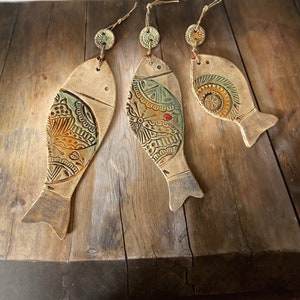 Puede incluir: Tres adornos de pescado de cerámica con intrincados patrones en tonos verdes, marrones y amarillos. Cada pez tiene un pequeño lazo en la parte superior para colgar.