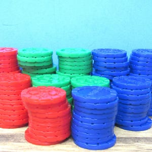 May include: A collection of plastic game tokens stacked in multiple columns. The tokens are circular with notched edges and are colored in bright red, green, and blue. Each token features a textured surface pattern on the top face.