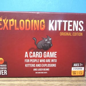 May include: The Exploding Kittens Original Edition card game box features a dark red background with a cartoon black cat holding a bomb. Text includes A CARD GAME FOR PEOPLE WHO ARE INTO KITTENS AND EXPLOSIONS AND LASER BEAMS AND SOMETIMES GOATS.
