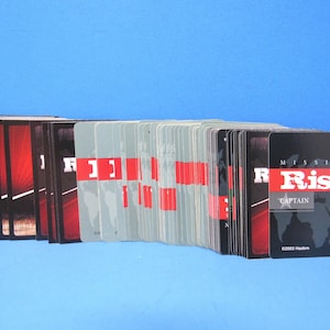 May include: A deck of "Mission: Risk" game cards fanned out against a blue background. The cards have a black and red design with the game title and the word "Captain" visible. The copyright date is 2003.