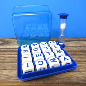 May include: A Boggle word game set featuring a blue plastic grid tray, sixteen white letter cubes with blue text, a clear blue plastic dome cover, and a blue-capped hourglass timer with white sand, all displayed as a complete tabletop game.
