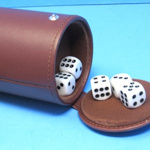 May include: A brown leather dice cup with a matching lid, containing four white dice with black dots. The dice are scattered on the lid and inside the cup. The cup is cylindrical and has a silver rivet.