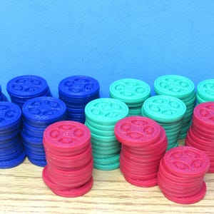 May include: A collection of circular plastic tokens stacked in multiple piles. The tokens are colored in blue, green, and red. Each token features a raised, embossed design on the top surface consisting of four small circular shapes.