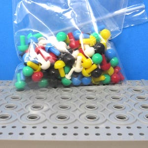 May include: A clear plastic resealable bag filled with a collection of small, mushroom-shaped plastic pegs in various colors including red, yellow, blue, green, white, and black, resting on a gray perforated pegboard base with circular holes.