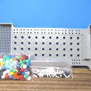 May include: A gray plastic Mastermind board game with a grid of circular holes and two storage compartments on the ends. Included are two clear plastic bags containing numerous small, round, multi-colored game pegs and smaller black and white pegs.