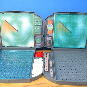 May include: Two gray plastic board game units featuring vertical radar screens with green circular displays and horizontal grids for gameplay. Each unit includes compartments filled with small red and white plastic game pieces and gray ship figures.