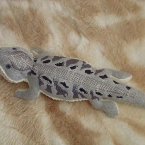 May include: A gray and white plush lizard with a glittery finish. The lizard has a long tail and a textured back.