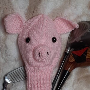 May include: A pink knitted golf club head cover shaped like a pig, with black eyes and a snout. The cover is placed over a golf club, with other clubs visible in the background. The cover is made of soft, textured yarn.
