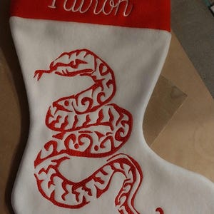 Peut inclure: Chaussette de Noël blanche avec un revers rouge et un motif de serpent brodé en rouge. Le revers est brodé du nom "Patrón" en blanc.