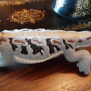 Puede incluir: Un juguete de peluche de un dragón barbudo, con un cuerpo gris claro, marcas marrones y negras y una cabeza dorada clara. El juguete está tumbado sobre una superficie de madera.