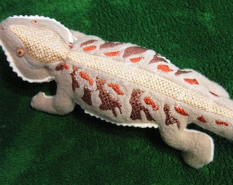 May include: A beige plush toy lizard with brown and orange embroidered details. The lizard has a textured, fuzzy surface and a stitched-on mouth.