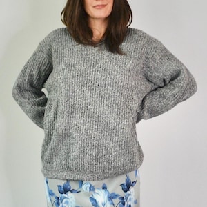 May include: A gray, ribbed knit sweater with a V-neck. The sweater has long sleeves and a relaxed fit. The person is wearing a floral print skirt. The sweater is a neutral color and could be worn in the fall or winter.