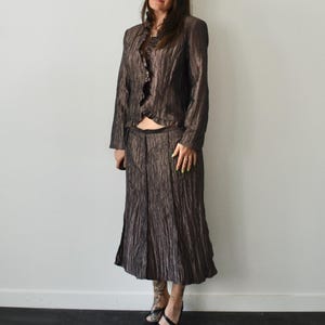 May include: A brown, textured skirt suit. The suit includes a long-sleeved jacket with a ruffled front and a matching midi skirt. The fabric has a subtle sheen, and the outfit appears to be vintage.