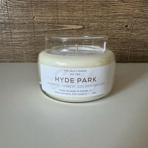 May include: A clear glass jar candle labeled "The Hyde Park" with the text "Jasmine, Amber, Golden Woods." The candle is hand-poured in Tampa, FL, and is a 100% natural soy candle. The candle is 11oz.