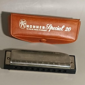 May include: A Hohner Special 20 harmonica with a brown leather case. The harmonica is silver and black, with the numbers 1 through 10 visible. The case has the Hohner logo and text that reads "Special 20".