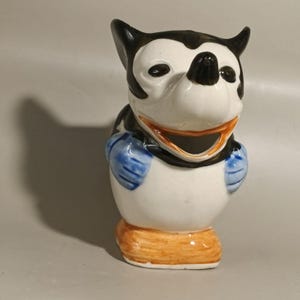 Puede incluir: Figura de cerámica de un perro de dibujos animados con orejas negras, nariz negra y cara blanca. El perro tiene guantes azules y una base naranja. Artículo decorativo.