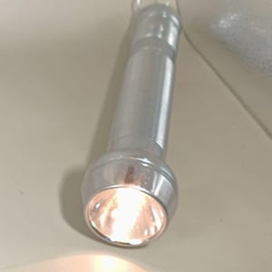 May include: A silver flashlight with a bright light emanating from the lens. The flashlight has a ribbed handle and a loop at the top. The light is a warm yellow color.