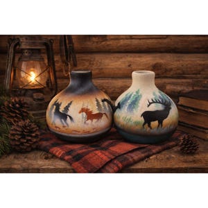 May include: Two ceramic vases with hand-painted nature scenes. One vase features running horses, the other a silhouette of a deer. The vases have a rustic, earthy color palette, with a dark gradient at the top.