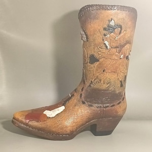 May include: A decorative brown cowboy boot with a detailed relief carving of a Native American on horseback. The boot features a pointed toe, a small heel, and a textured surface. The boot is a unique piece of Western-themed decor.