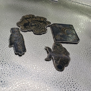 Può includere: Una collezione di toppe grigio scuro con dettagli color oro. Le toppe presentano varie forme, tra cui una mano, un libro, un fiore e un disegno circolare con testo. Le toppe sembrano ricamate o applicate.