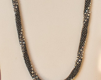 May include: A beaded necklace with a dark gray base and scattered iridescent beads. The necklace is draped against a neutral background, showcasing its texture and design. The beads are small and tightly packed, creating a rope-like appearance.