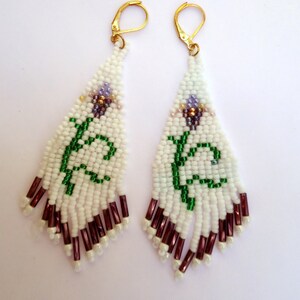 May include: A pair of beaded earrings with a white background. The earrings are white with a green vine design and purple and brown accents. The bottom of the earrings have dark red beads. The earrings have gold-colored hooks.
