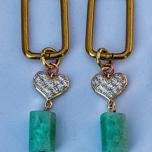 May include: A pair of gold-colored rectangular hoop earrings. Each earring features a heart-shaped charm encrusted with small, clear crystals and a rectangular, light green stone pendant. The earrings have a delicate, elegant design.