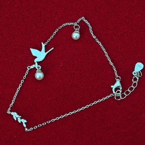 May include: A silver chain bracelet with a delicate bird charm and two pearl accents. The bracelet has a lobster clasp closure.
