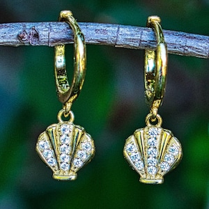 May include: A pair of gold-colored hoop earrings with seashell pendants. Each pendant is encrusted with small, clear gemstones. The earrings are hanging from a small, brown branch against a blurred green background.