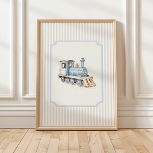 May include: A framed watercolor illustration of a blue toy train with a white background. The train has a black chimney and wheels. The frame has a light brown wood finish and a white and light blue striped background.