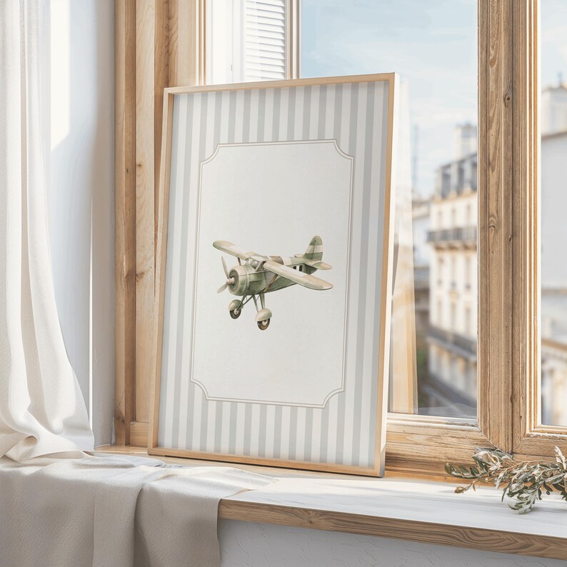 May include: A framed print featuring a watercolor illustration of a vintage airplane. The artwork is set against a backdrop of vertical gray and white stripes, framed in light wood. The print is displayed on a windowsill.
