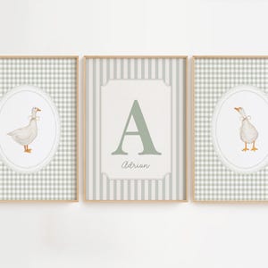 May include: Three framed wall art prints with light wooden frames. The two outer prints feature a white goose in an oval frame against a sage green gingham background. The center print displays a large letter A and the name Adrian in script.