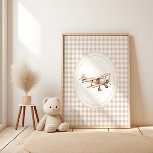 Pu&ograve; includere: Una stampa incorniciata con un'illustrazione ad acquerello di un biplano vintage. L'opera d'arte &egrave; ambientata su uno sfondo a scacchi beige e bianco all'interno di una cornice ovale. La stampa &egrave; esposta in una cornice di legno chiaro, con un orsacchiotto e una pianta decorativa nelle vicinanze.