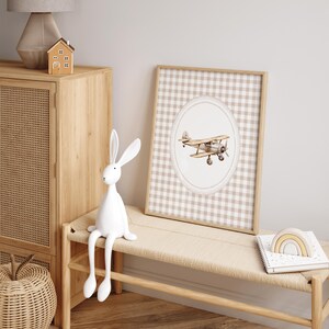 Pu&ograve; includere: Una stampa incorniciata con un'illustrazione ad acquerello di un biplano vintage. L'opera d'arte &egrave; ambientata su un motivo a quadretti beige e bianco, incorniciata in legno chiaro. La scena include una statuina di coniglio bianco e una panca di legno.